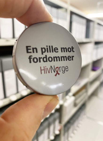 En pilleboks holdes mot en bakgrunn av arkivhyller. På lokket står: «En pille mot fordommer, Hiv Norge.»