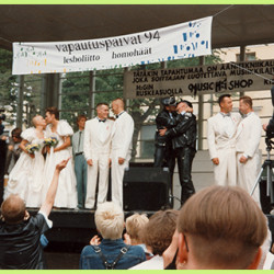 Four same-sex couples on stage. First on the left is a lesbian couple kissing, with matching white wedding dresses. The second couple is a gay couple with matching white suits. The third one is a gay couple in leather, kissing. The fourth one is a gay couple in matching suits, looking at each other. Above them is a banderoll with the name of the the event: Vapautuspäivät 1994, lesboliitto, homohäät - lesbian marriage, gay wedding.