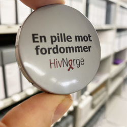 En pilleboks holdes mot en bakgrunn av arkivhyller. På lokket står: «En pille mot fordommer, Hiv Norge.»